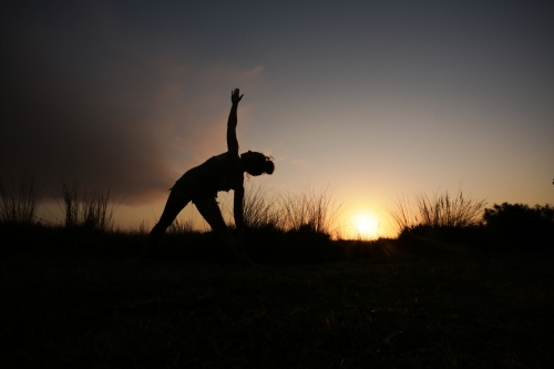 Silhouette shot of a young woman doing exercise on a field with grass under the sun set light - Australian Stock Image