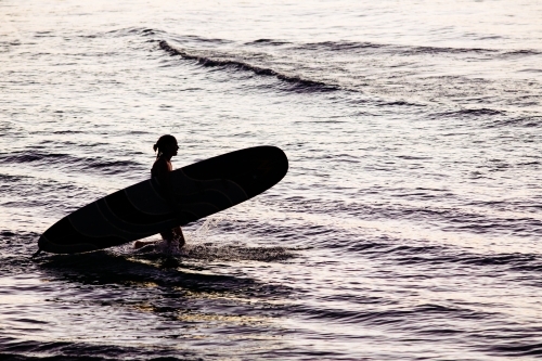 Silhouetted female surfer holding a longboard entering the water at dusk, Byron Bay. - Australian Stock Image