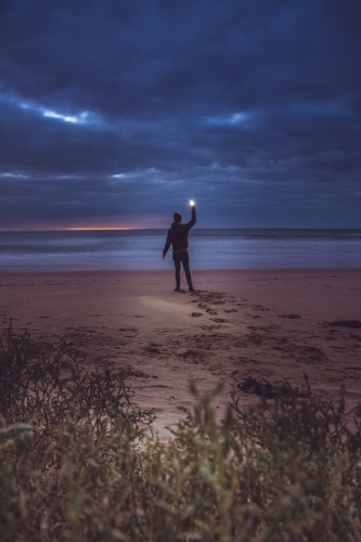 Silhouetted Figure Holding Up a Light on a Dark Beach - Australian Stock Image