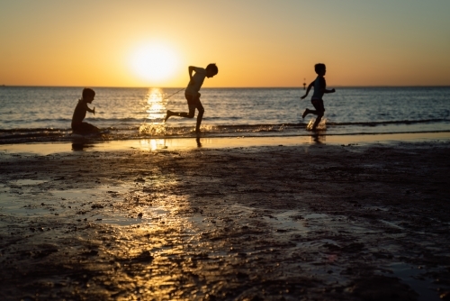Silhouettes of three children playing on the beach at sunset - Australian Stock Image