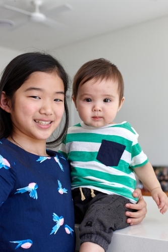 Sister holding younger baby brother in kitchen - Australian Stock Image