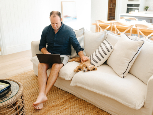 Small dog lying next to a man holding a laptop while sitting on the sofa. - Australian Stock Image