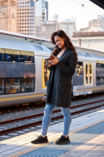 Smiling Asian woman waiting at train station with mobile phone - Australian Stock Image