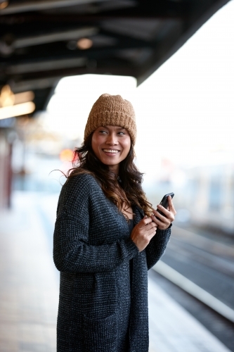 Smiling Asian woman waiting at train station with mobile phone - Australian Stock Image