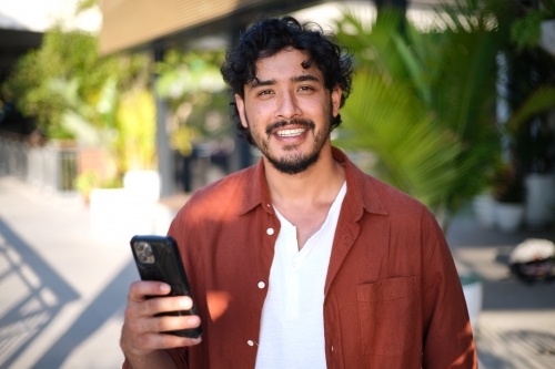 Smiling curly haired man with brown shirt and white tshirt holding his phone - Australian Stock Image