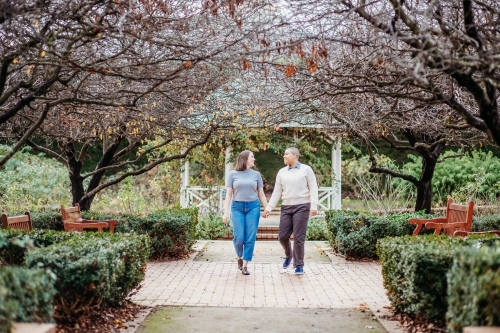 smiling lgbt  couple holding hands with dead trees, shrubs and wooden benches on the side - Australian Stock Image