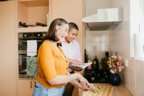 Smiling lgbtqi couple  with one chopping leeks and the other reading a book - Australian Stock Image