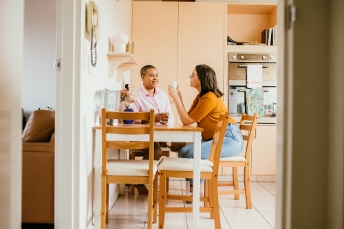 Smiling lgbtqi  couple sitting on a kitchen table drinking on a white mug - Australian Stock Image