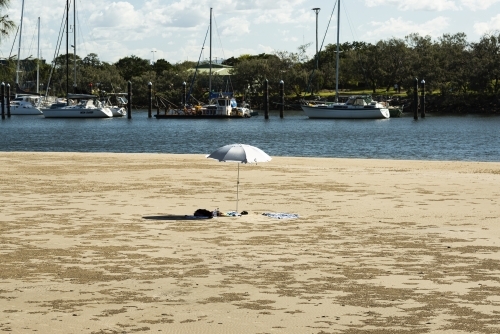 Solitary solitary beach umbrella in beach scene with boats moored in background at riverside - Australian Stock Image