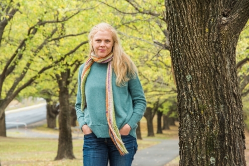 Strawberry blonde woman standing next to a tree in winter with hands in pockets.