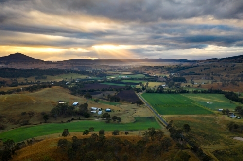 Sun rays piercing through the cloudy sky over farmland rimmed in hills - Australian Stock Image