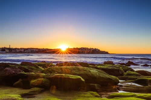 Sun rising over moss covered rocks along coastline with blue sky - Australian Stock Image