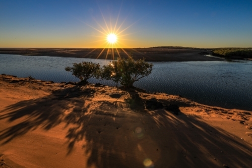 Sun rising through blue sky over ocean channel and sandy coastline - Australian Stock Image