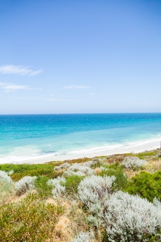 Sunlit coastland with view of shrubs, beach and clear blue coastal waters - Australian Stock Image