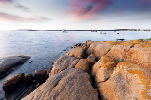 Sunset over South Australian fishing harbour, lichen covered granite rocks and boats - Australian Stock Image