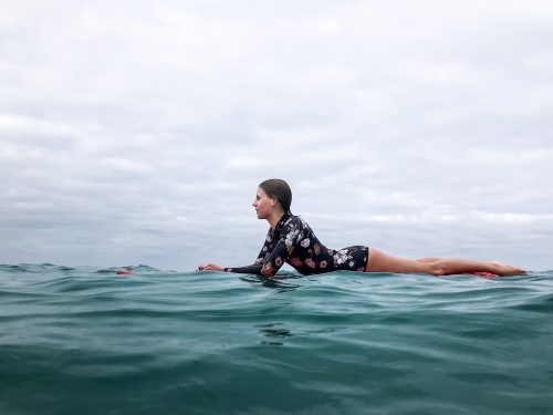 Surfing Teenage girl lying on red surfboard in ocean wearing floral swimsuit on overcast day - Australian Stock Image