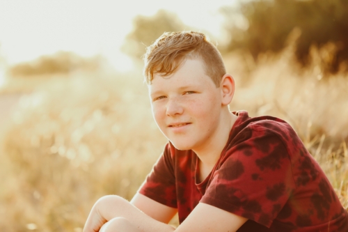 Teen boy sitting in long dry grass in golden afternoon light - Australian Stock Image