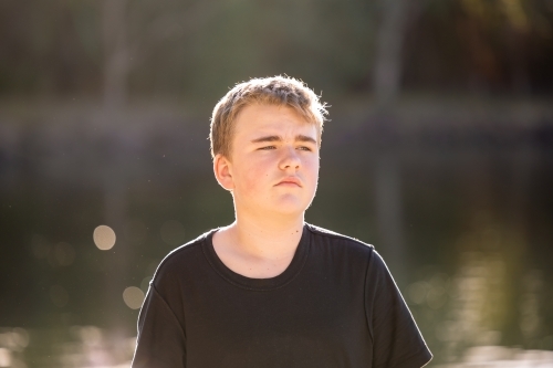 Teen boy standing in golden afternoon light by the water on holiday - Australian Stock Image