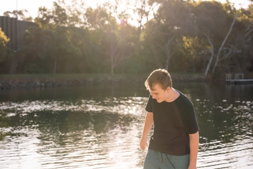 Teen boy standing in golden afternoon light by the water on holiday - Australian Stock Image