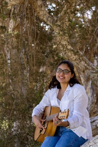 teen girl in white shirt wearing glasses strumming guitar sitting on a tree - Australian Stock Image