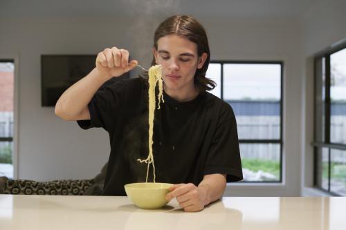 Teenage boy eating noodles at a kitchen bench