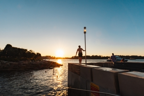 Teenage boy standing on ledge by water in bright setting sun - Australian Stock Image