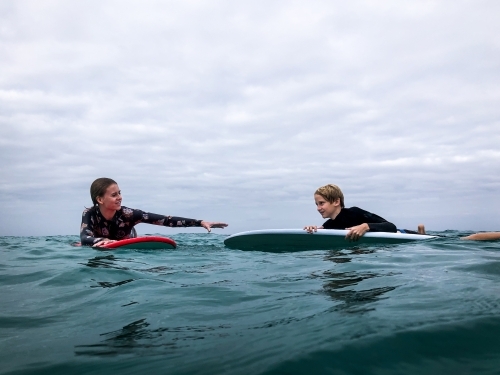 Teenage girl assisting teenage boy in the surf on longboard surfboards on overcast day - Australian Stock Image