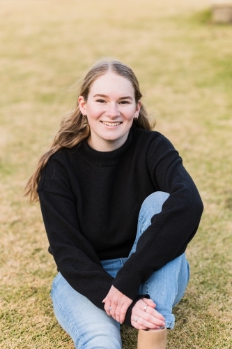 Teenage girl sitting on grass smiling with arm leaning on leg - Australian Stock Image