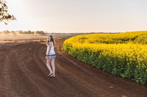 Teenager walking on dirt road on farm looking over shoulder next to paddock of canola - Australian Stock Image