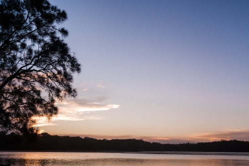 The sun sets behind the silhouette of a tree on the left overlooking a large, smooth lake. - Australian Stock Image