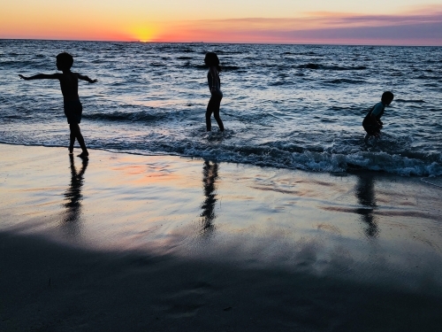 Three children playing on oceans edge with sun setting in background - Australian Stock Image