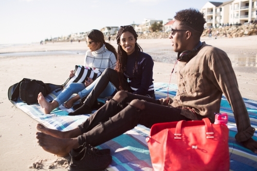 Three friends sitting on a picnic blanket relaxing at the beach - Australian Stock Image