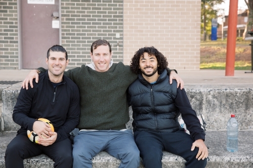 Three mates sitting on  soccer clubhouse  steps, with one man holding a soccer ball - Australian Stock Image