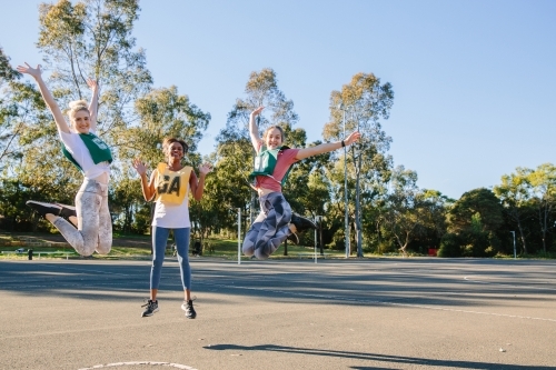 three young women jumping in mid air on a sunny day with trees at the back - Australian Stock Image