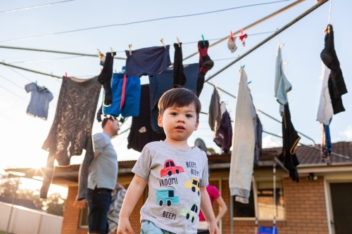 Toddler boy in backyard playing while parents get in washing from hills hoist line - Australian Stock Image