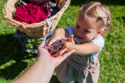 Toddler girl handing pegs to mum to help with hanging out washing in backyard - Australian Stock Image