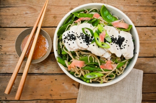 Top shot of a bowl of vegetables with noodles and cut chicken with brown chopsticks and orange sauce - Australian Stock Image