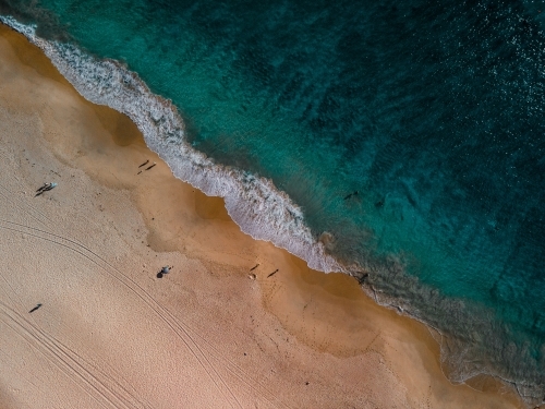 Top view shot of a beach with a white shore and blue waters with a few people standing on the shore - Australian Stock Image