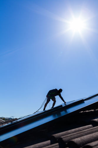Tradie man laying solar panel on roof under blue sky during solar power system installation job - Australian Stock Image