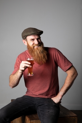 Trendy man with ginger beard and flat cap, holding a glass of beer - Australian Stock Image