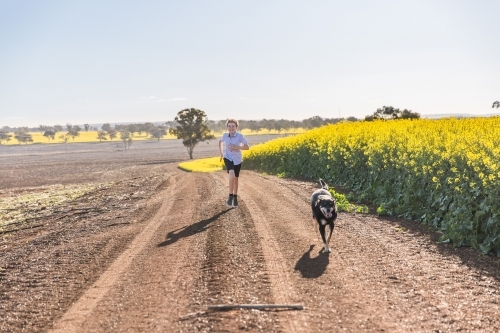 Tween boy having fun chasing kelpie dog on dirt road on farm near canola paddock