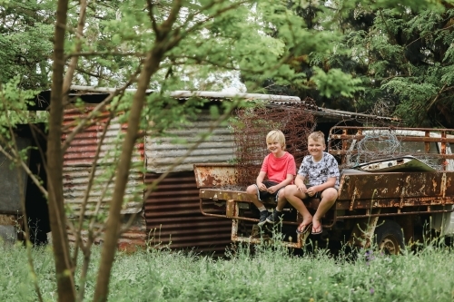 Two boys sitting on ruins of old ute on abandoned farm. Urban exploration, run down property - Australian Stock Image