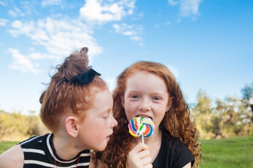 Two girls outside eating a rainbow lollipop - Australian Stock Image