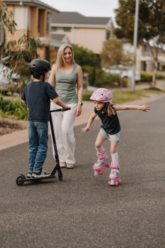 Two kids on roller skates and scooter on the street with mum. - Australian Stock Image