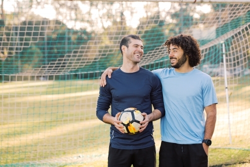 Two men looking at each other and laughing while standing in a soccer goal, holding a ball - Australian Stock Image