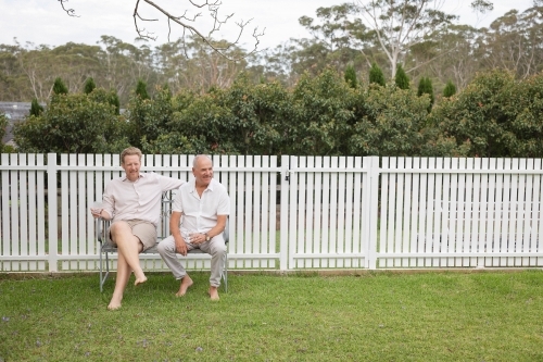 Two men sitting on backyard bench against white fence - Australian Stock Image