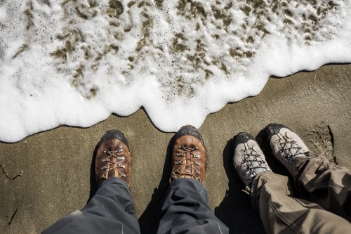 Two pairs of hiking boots on sandy beach with whitewash - Australian Stock Image