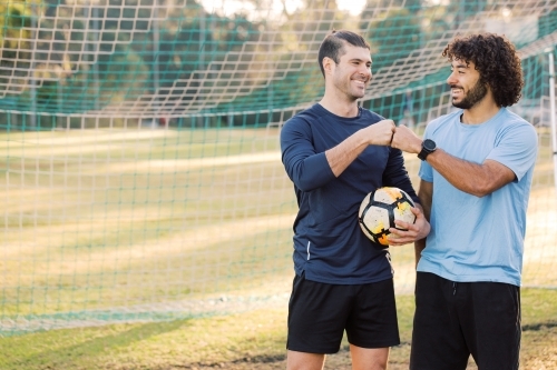 Two smiling young men standing on the field fist bumping near the net with a soccer ball - Australian Stock Image