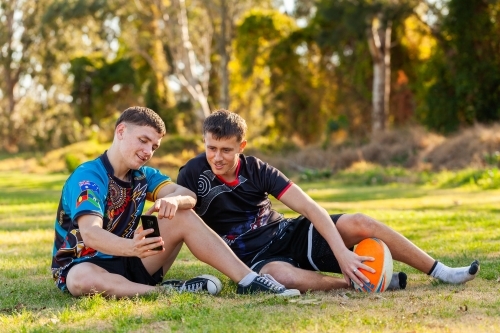 Two teen Aboriginal sport players chatting together at the edge of sports field looking at phone - Australian Stock Image