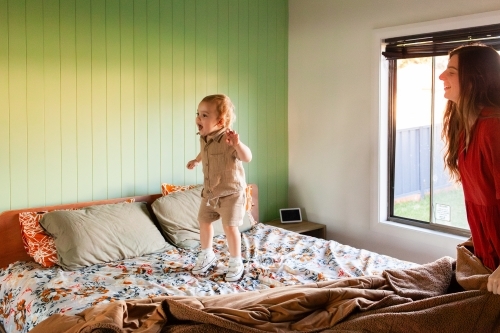 Two year old toddler jumping on parents bed with mum smiling - Australian Stock Image